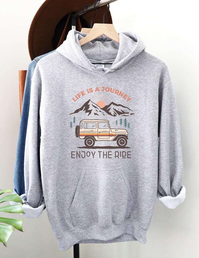 May include: Grey hooded sweatshirt with a graphic of a vintage jeep driving through a mountain landscape. The text "Life is a Journey, Enjoy the Ride" is printed on the sweatshirt.