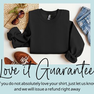 May include: A black sweatshirt laid out on a white surface with a pair of brown shoes, sunglasses, and a straw hat. The text "Love it Guarantee" is written below the sweatshirt. The text below the guarantee reads "If you do not absolutely love your shirt, just let us know and we will issue a refund right away."