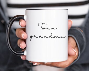 Może przedstawiać: Biały kubek ceramiczny z czarnym uchwytem i czarnym rantem. Kubek ma napis "Twin grandma" napisany czarnym kursywą.