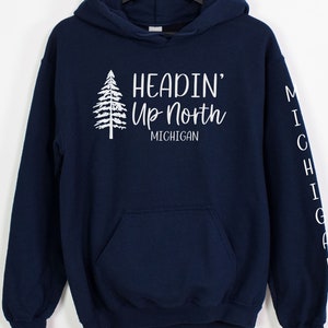 May include: Navy blue hooded sweatshirt with a white graphic of a pine tree and the text "Headin' Up North Michigan". The left sleeve has the word "Michigan" vertically down the sleeve.