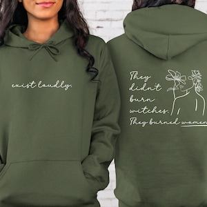 May include: An olive green hoodie with the text "exist loudly" on the front. The back of the hoodie features the text "They didn't burn witches. They burned women" with a line drawing of a woman's face and flowers.