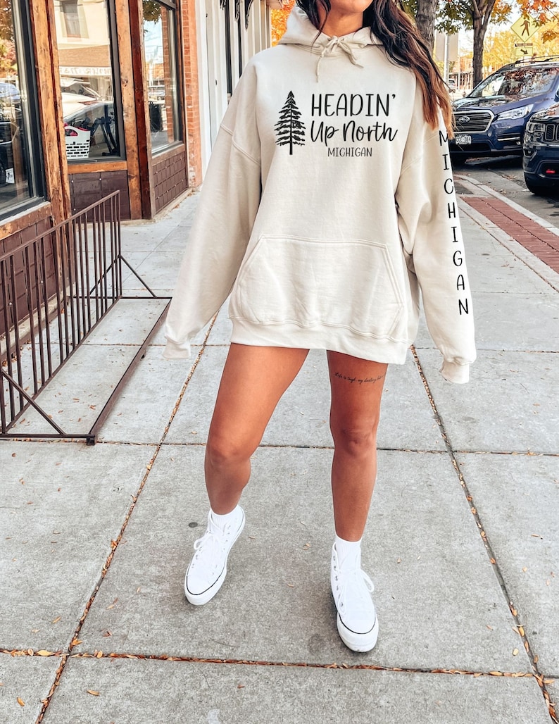 May include: A cream-colored hoodie with a black pine tree graphic and the text "Headin' Up North Michigan". The left sleeve has the word "Michigan" vertically down the side.