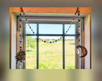 May include: A decorative suncatcher featuring a chain of multicoloured faceted beads draped across a window frame. The chain connects a metal sun charm on the left and a metal crescent moon charm on the right, each with a clear crystal drop pendant.