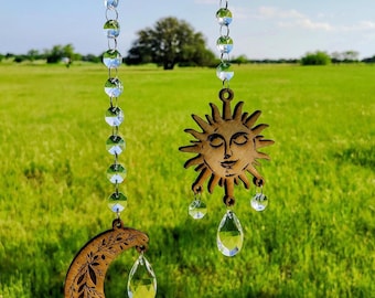 May include: Wooden sun and moon hanging ornaments with crystal accents. The sun has a smiling face and the moon has a floral design. Both are suspended by clear crystal bead strands. The background is a green field under a blue sky.