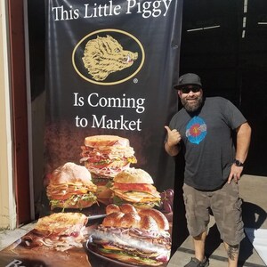 May include: A black and white banner with the text "This Little Piggy Is Coming to Market" and a cartoon boar's head. The banner is next to a man wearing a black t-shirt, khaki shorts, and a black baseball cap. The banner also features a photo of several Boar's Head deli sandwiches.