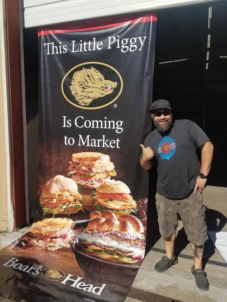 May include: A black and white banner with the text "This Little Piggy Is Coming to Market" and a cartoon boar's head. The banner is next to a man wearing a black t-shirt, khaki shorts, and a black baseball cap. The banner also features a photo of several Boar's Head deli sandwiches.