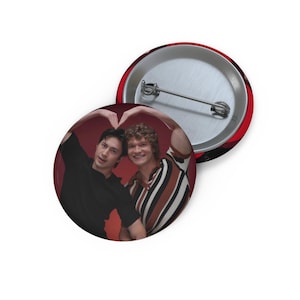 Peut inclure: Un badge rond à épingle présentant une photographie de deux personnes formant un cœur avec leurs bras au-dessus de leur tête. L'une porte un t-shirt noir, tandis que l'autre porte une chemise à rayures marron, noires et blanches.