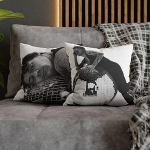 May include: Two decorative throw pillows with black and white images. One pillow features a person lying on a disco ball, and the other shows a person leaning on a disco ball. The pillows are on a gray couch.