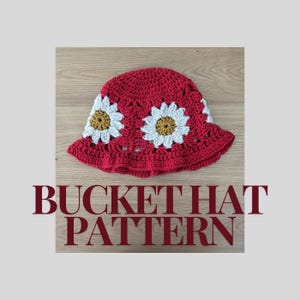 May include: A red crochet bucket hat with white and yellow daisy flowers. The hat is on a wooden surface. BUCKET HAT PATTERN is written in red letters below the hat.