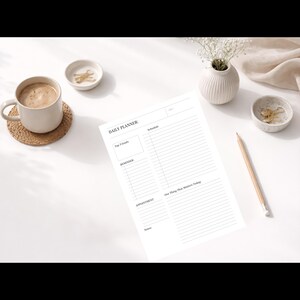 Op de afbeelding: Een witte dagplanner met de tekst "DAILY PLANNER" en secties voor doelen, herinneringen, afspraken en notities. Een kopje koffie, een potlood en decoratieve kommen staan ook op het witte oppervlak.