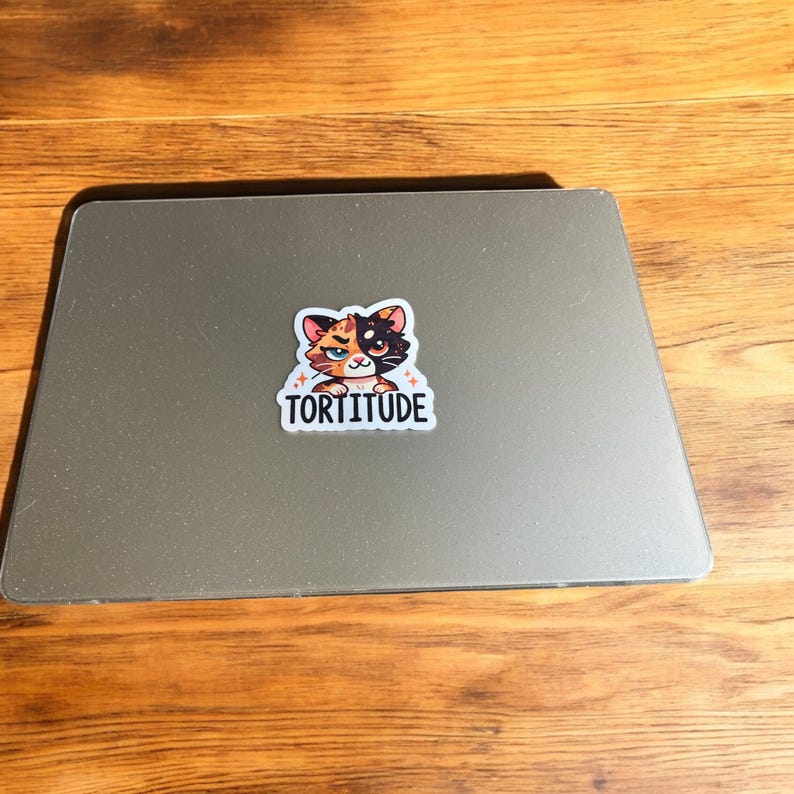 Op de afbeelding: Een witte sticker met een cartoonkat met oranje, zwart en wit bont. De kat heeft een brutale uitdrukking en is omringd door kleine witte sterren. De tekst "Tortitude" is onder de kat gedrukt.