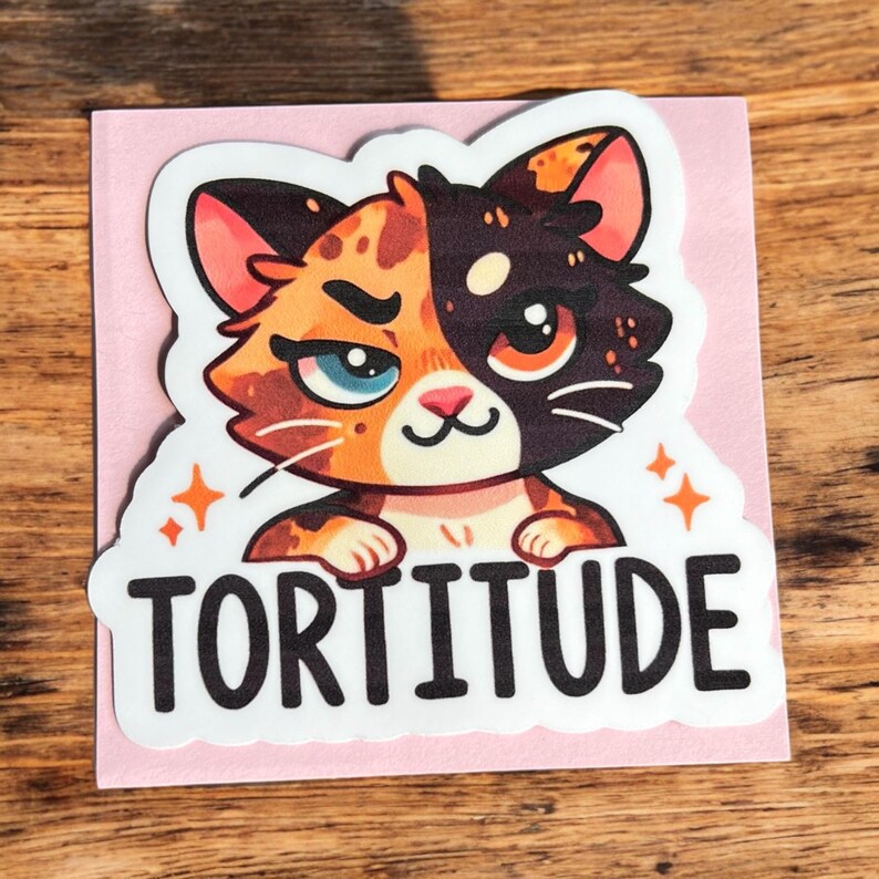 Op de afbeelding: Een sticker met een cartoonkat met oranje, zwart en wit bont. De kat heeft een chagrijnige uitdrukking en kijkt recht naar de kijker. De tekst "TORTITUDE" is onder de kat gedrukt.