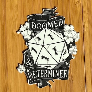 May include: Black and white sticker with a stylized illustration of a twenty-sided die with the words "Doomed & Determined" on a banner.