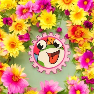May include: A pink cog with a cartoon frog face in the centre. The cog is surrounded by a circle of pink, yellow and orange flowers.