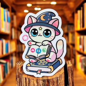 Puede incluir: Un gato blanco con un sombrero de bruja azul y blanco con un diseño de estrella y luna creciente. El gato sostiene un libro rosa, azul y verde con un pentagrama en la portada. El gato está sentado en una pila de libros con un pentagrama en el libro superior.