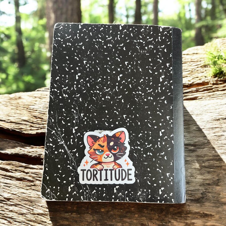 Op de afbeelding: Een zwart-wit compositieboek met een sticker van een cartoonkat met oranje, zwart en wit bont. De sticker zegt "Tortitude" in witte letters.