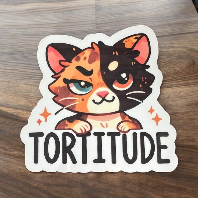 Op de afbeelding: Een witte sticker met een cartoonillustratie van een kat met oranje, zwart en wit bont. De kat kijkt de kijker aan met een ondeugende uitdrukking. De tekst "TORTITUDE" is in zwart onder de kat gedrukt.
