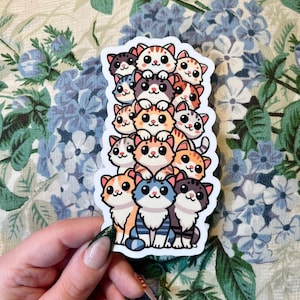 May include: A sticker featuring a stack of 10 cartoon cats with different colored fur and patterns. The cats are all looking at the viewer with big eyes and smiling. The sticker is on a white background with a floral pattern.