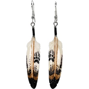 May include: A pair of dangle earrings featuring carved bone feathers with black bear paw prints. The feathers are white with brown and black accents. The earrings have silver hooks.