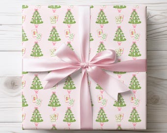 以下が含まれることがあります： 緑のクリスマスツリーとトランプの繰り返し模様が描かれたピンク色の包装紙。明るいピンク色のサテンリボンが、包まれたギフトの周りにリボン結びされています。背景は白い木製の表面です。