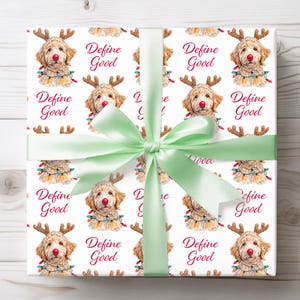 May include: A square gift box wrapped in white paper featuring a repeating pattern of a golden doodle dog wearing reindeer antlers, a red nose, and colorful Christmas lights. The text Define Good is printed in red. A light green ribbon is tied in a bow.