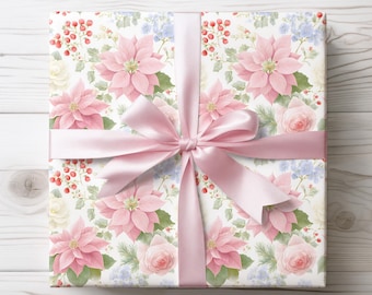 May include: A square gift box wrapped in white paper featuring a floral pattern of pink poinsettias, pale pink roses, small blue flowers, green leaves, and red berries. A wide, smooth, light pink satin ribbon is tied in a bow around the center.
