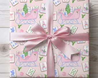 May include: A square gift box wrapped in light pink paper featuring a repeating pattern of pink sleighs, Christmas trees, mahjong tiles, and holly leaves, tied with a wide, smooth, light pink satin ribbon centered in a large bow.