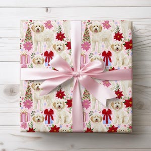 May include: A square gift box wrapped in light pink paper featuring a repeating pattern of cream-colored doodle dogs, green Christmas trees, red poinsettias, and blue gift boxes with red bows, tied with a wide, smooth, light pink satin ribbon.