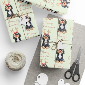 May include: Gift-wrapped presents with light green paper featuring a repeating pattern of a black and brown dog wearing a red bow and antlers. The words "Merry Christmas" are printed in red. The gifts are tied with twine and adorned with white star-shaped tags.