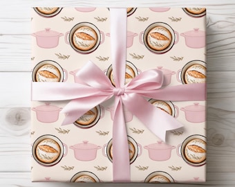 Könnte beinhalten: Ein Geschenk, das mit einem hellrosa Satinband verpackt ist. Das Geschenkpapier zeigt ein sich wiederholendes Muster aus rosa Kochtöpfen, Brotlaiben und Zweigen auf cremefarbenem Hintergrund. Das Geschenk liegt auf einer weißen Holzoberfläche.