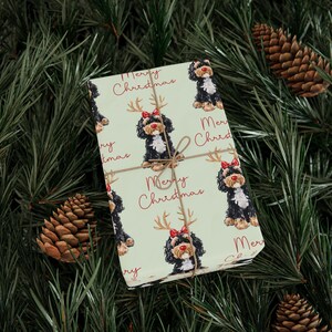 May include: A wrapped gift with a light green background featuring a repeating pattern of a black and white dog wearing reindeer antlers and a red bow. The words "Merry Christmas" are written in red. The gift is tied with twine.