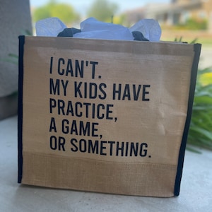 May include: A tan jute tote bag with black trim and handles. The bag features the text "I CAN'T. MY KIDS HAVE PRACTICE, A GAME, OR SOMETHING." in black block letters. White tissue paper is visible inside the bag.