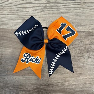 May include: A blue and orange cheer bow with a baseball stitch design. The bow has the name "Ricci" and the number "17" on it.