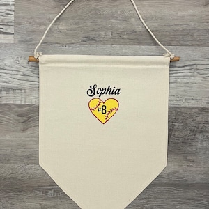 May include: Cream-colored pennant banner with a wooden dowel and hanging cord. The banner features the name "Sophia" in black script above a yellow heart with a softball design and the number "8".