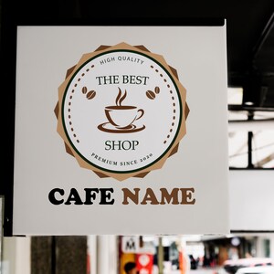 May include: A white sign with brown and black text and a brown and white logo. The logo features a coffee cup with steam coming from it and the text "The Best Shop Premium Since 2020". The text below the logo reads "Cafe Name".