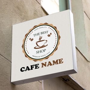 May include: A white sign with brown and green accents advertising a cafe. The sign has a logo with a coffee cup and the text "The Best Shop" and "Premium Since 2020". The text "Cafe Name" is in large, bold letters below the logo.