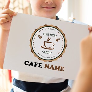 May include: A white sign with a brown logo featuring a coffee cup and the text "The Best Shop" in a brown circle with the text "High Quality" and "Premium Since 2020" in smaller text. The text "Cafe Name" is below the logo in brown.