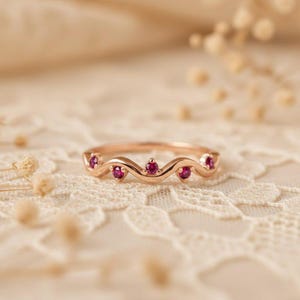 May include: A rose gold ring featuring a wavy design, adorned with five small, round, red gemstones. The ring is presented on a white lace surface, with a soft, blurred background.