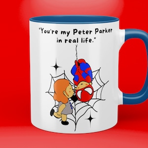 Puede incluir: Taza de cer&aacute;mica blanca con borde y asa azules. La taza presenta una imagen de dibujos animados de Spider-Man colgando boca abajo de una telara&ntilde;a con una ni&ntilde;a bes&aacute;ndolo. El texto en la taza dice "You're my Peter Parker in real life."