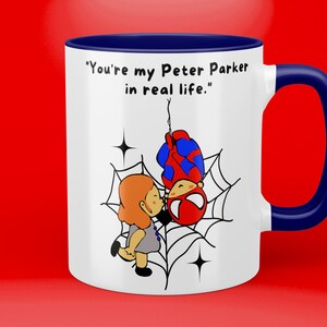 Puede incluir: Taza de cer&aacute;mica blanca con borde y asa azules. La taza presenta una imagen de dibujos animados de Spider-Man colgando boca abajo de una telara&ntilde;a con una mujer bes&aacute;ndolo. El texto en la taza dice "You're my Peter Parker in real life."