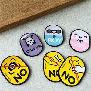 Könnte beinhalten: Eine Sammlung farbenfroher, ovaler Aufkleber. Designs umfassen einen Totenkopf mit "DANGER", ein lächelndes Gesicht mit Sonnenbrille und "BOY", ein lächelndes Gesicht mit Schleife und "GIRL" sowie gelbe Aufkleber mit "NO" und Verbotssymbolen.