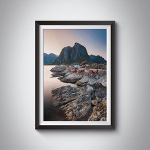 May include: A scenic photograph of a small village nestled between rocky mountains and a body of water. The village is made up of red-roofed houses and is surrounded by a rocky shoreline. The sky is a soft blue with a hint of pink in the distance.