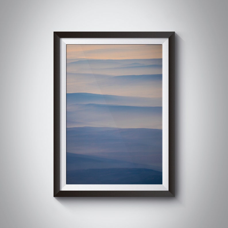May include: A framed landscape photograph featuring layers of blue and purple mountains partially obscured by a soft, hazy atmosphere. The sky transitions from a pale orange to a light blue, creating a serene and atmospheric scene.