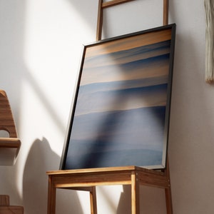 May include: A framed abstract art print featuring horizontal bands of blue and tan hues. The artwork is displayed on a wooden chair, leaning against a small wooden ladder. The scene is illuminated by natural light.