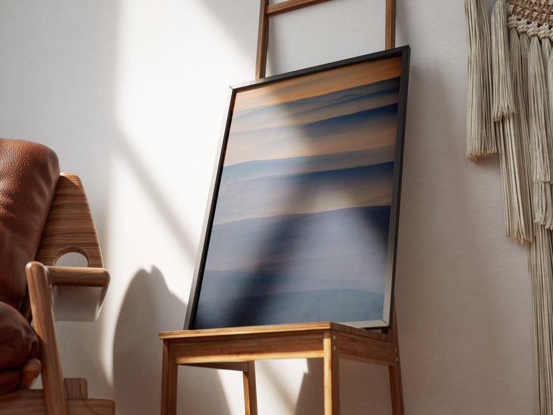 May include: A framed abstract art print featuring horizontal bands of blue and tan hues. The artwork is displayed on a wooden chair, leaning against a small wooden ladder. The scene is illuminated by natural light.