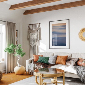 May include: A cozy living room with a neutral-toned sofa adorned with colorful throw pillows and a faux fur throw. A round coffee table with a glass top sits in front of the sofa. The room features a macrame wall hanging, framed artwork, and decorative mirrors.