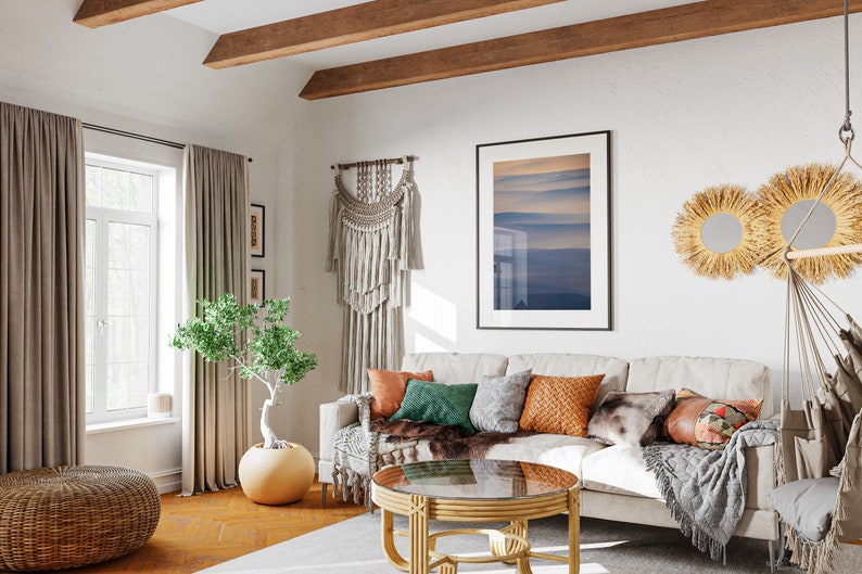May include: A cozy living room with a neutral-toned sofa adorned with colorful throw pillows and a faux fur throw. A round coffee table with a glass top sits in front of the sofa. The room features a macrame wall hanging, framed artwork, and decorative mirrors.
