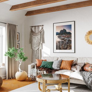 May include: A white couch with a variety of throw pillows and blankets in a living room with a large window, a wooden coffee table, and a macrame wall hanging.