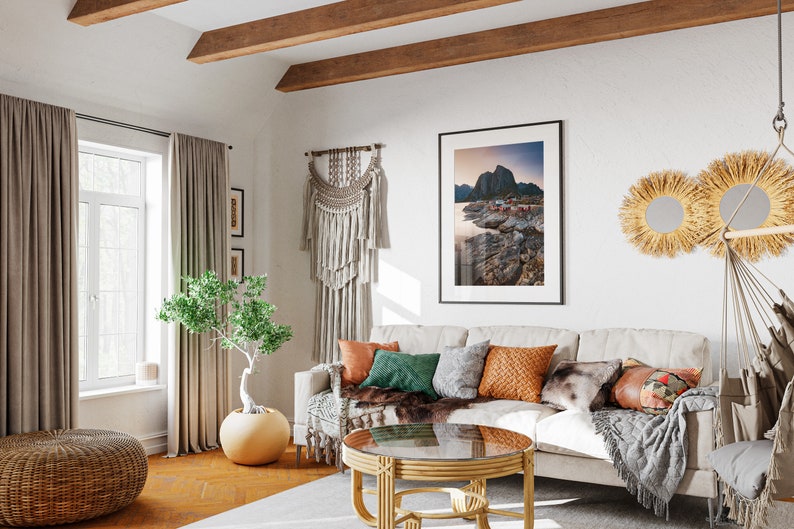May include: A white couch with a variety of throw pillows and blankets in a living room with a large window, a wooden coffee table, and a macrame wall hanging.