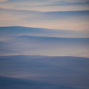 May include: An aerial view of layered mountain ranges shrouded in a soft, blue-gray mist. The sky transitions from a pale orange at the top to a deeper blue, creating a serene and atmospheric landscape.